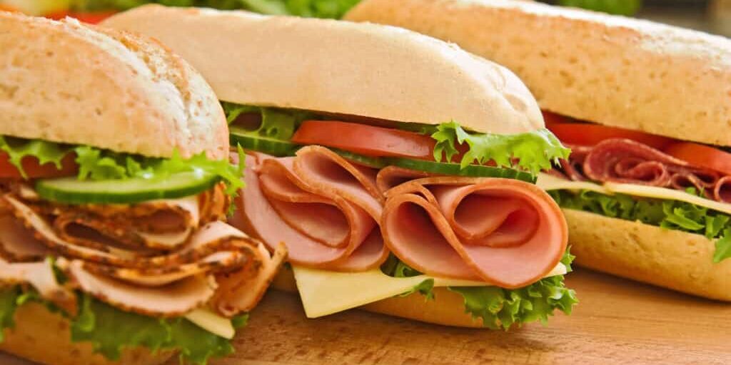 Three fresh sub sandwiches - turkey/chicken breast, ham &amp; swiss and salami on a cutting board. Focus on the ham sandwich